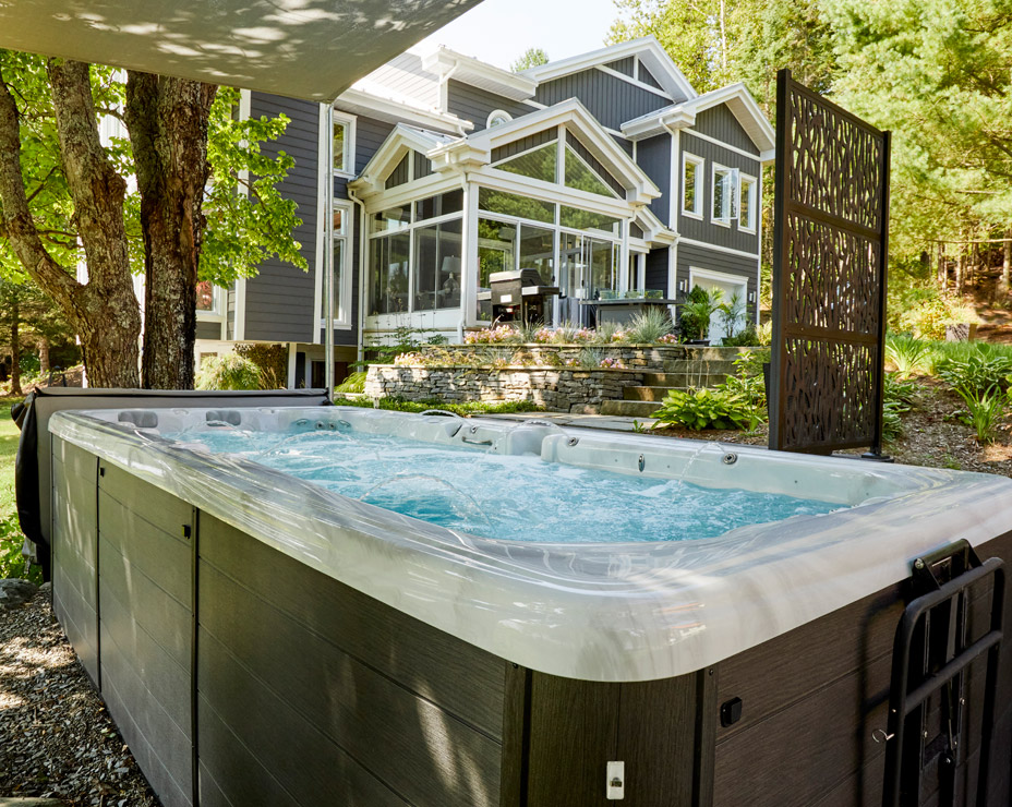 A Trevi Crossfit swim spa nestled in a backyard oasis. The spa is bubbling with clear water, positioned under a partial roof for shade. The scene showcases a lush green landscape, a modern house with a sunroom, and a decorative privacy screen.