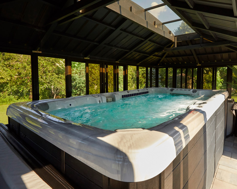 A Trevi exercise swim spa installed in a luxurious gazebo. Sunlight streams through the glass roof panels, illuminating the bubbling blue water. The spa is surrounded by dark wood paneling and overlooks a green backyard with trees.