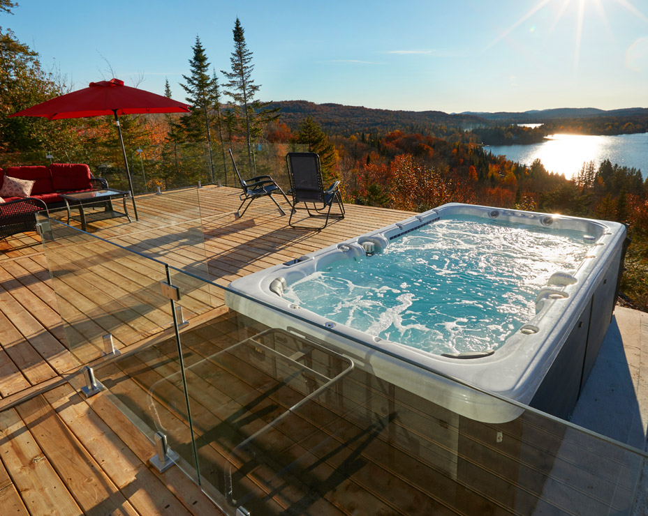 A luxurious outdoor deck with a Trevi exercise swim spa overlooking a breathtaking autumn landscape. The spa is bubbling invitingly, surrounded by lounge chairs and a cozy seating area. A glass railing offers unobstructed views of the colorful fall foliage and a serene lake in the distance.