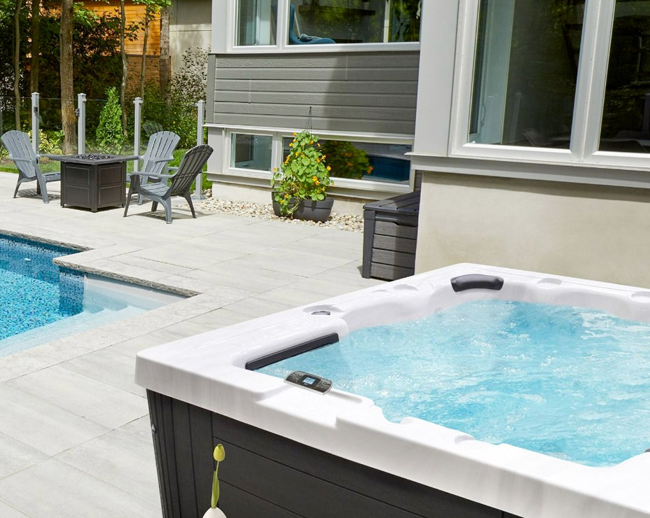 A Trevi L300 spa with steaming water, positioned on a patio beside an in-ground pool. The spa has a black exterior and is surrounded by gray patio stones. The backyard features a modern house, a fire pit with seating, and lush greenery.
