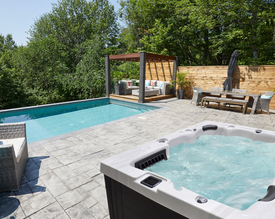 A luxurious backyard oasis featuring a Trevi L-400 hot tub on a stamped concrete patio. The spa is steaming, inviting relaxation, and is positioned near a modern in-ground pool.