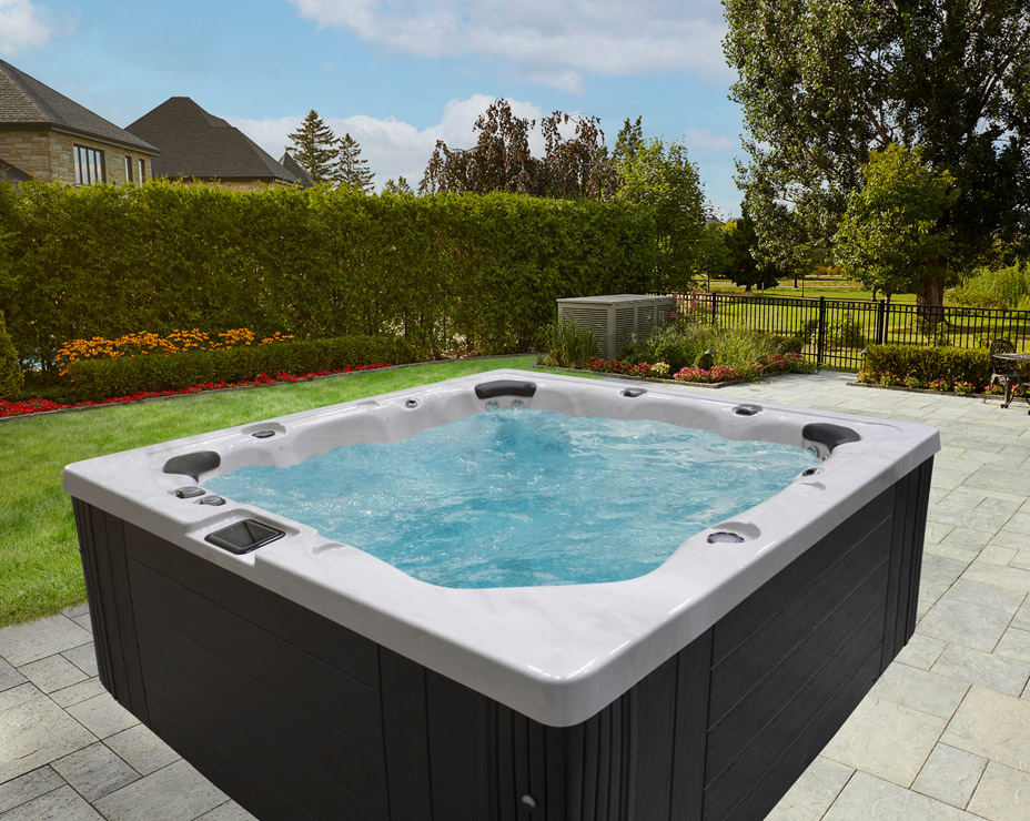 A Trevi L-450 hot tub sits on a paved patio in a sunny backyard. The spa has a black base and a white interior filled with clear, bubbling water.