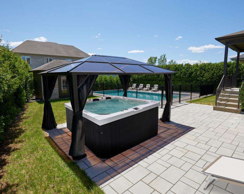 A luxurious backyard retreat featuring a Trevi L-950 hot tub nestled under a gazebo. The spa's steaming water invites relaxation, while the adjacent in-ground pool offers a refreshing escape. The surrounding patio, landscaping, and clear blue sky create a perfect outdoor oasis.