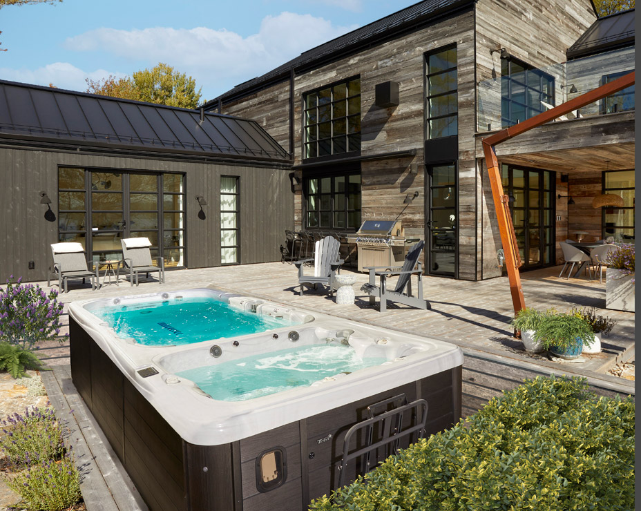 A luxurious backyard featuring a Trevi Spartan swim spa on a spacious wooden deck. The spa has two distinct sections: one with clear, calm water and another with bubbling jets. The modern, dark-colored spa complements the rustic wood and stone exterior of the house. Lounge chairs, an outdoor kitchen, and manicured landscaping create a serene outdoor living space.