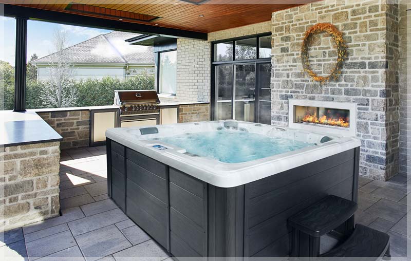 A luxurious Trevi Classik spa nestled in a covered outdoor patio. The spa features a sleek black exterior and inviting, steaming water. The surrounding space includes a stone fireplace, an outdoor kitchen, and comfortable seating, creating a perfect ambiance for relaxation and well-being.