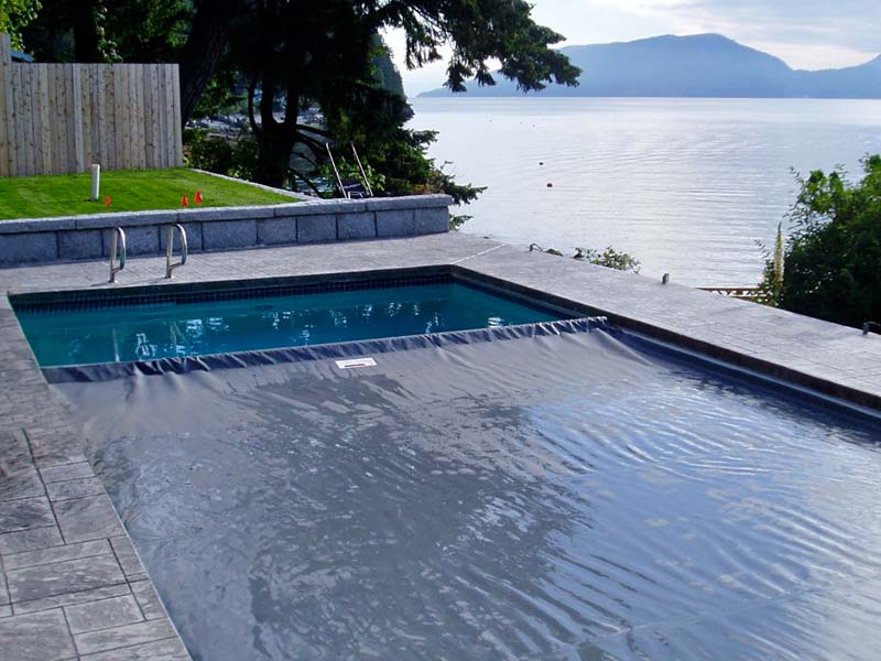 A rectangular pool with a Coverstar automatic pool cover partially closed. The pool is situated on a concrete patio with stunning views of a lake and mountains in the background.