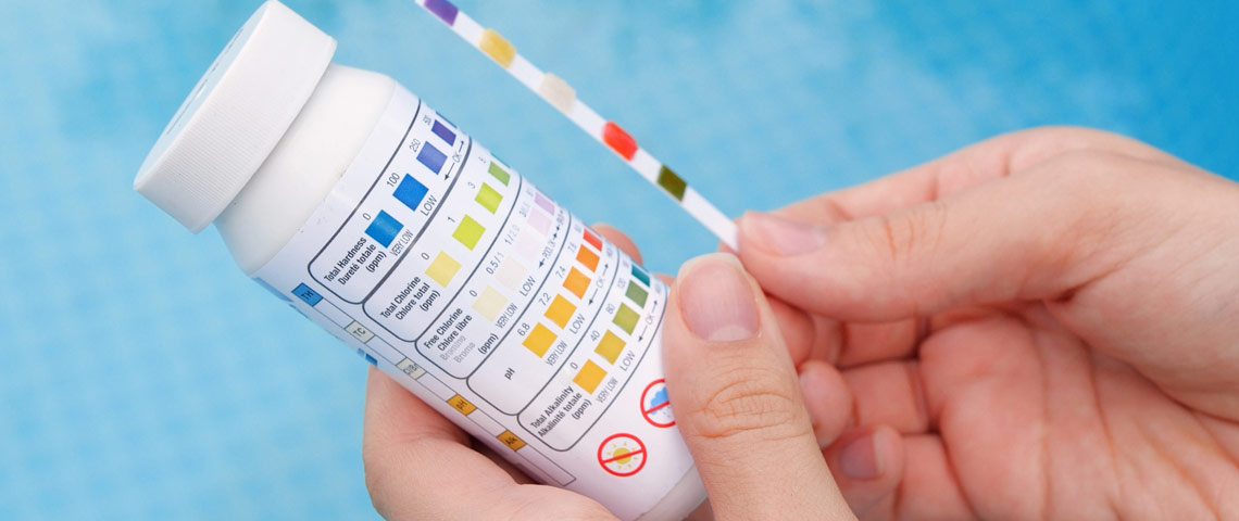 Close-up of a hand holding a pool test strip against a color comparison chart, illustrating the importance of testing water chemistry for pool maintenance.