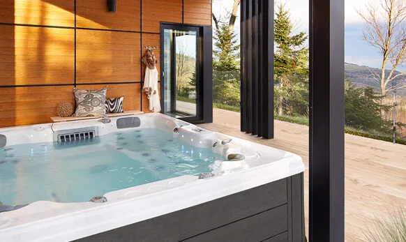 A square Trevi hot tub with steaming water is positioned on a wooden deck. The deck is attached to a modern house with large windows showcasing a scenic mountain view.